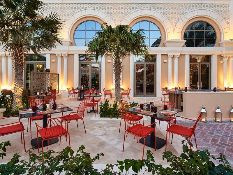 Hotel outdoor area with red chairs, tables, and palm trees at dusk.