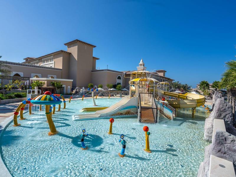 Outdoor water playground with slides, fountains, and shallow pool at a resort under clear sky.