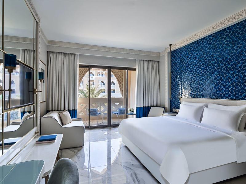 Modern hotel room with large bed, blue accent wall, and balcony overlooking palm trees.