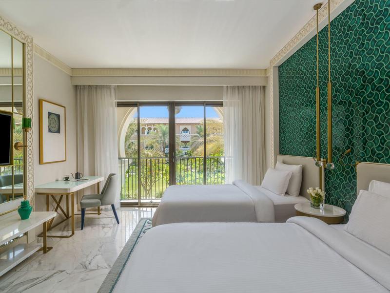 Modern hotel room with two beds, green patterned accent wall, desk, and balcony view.