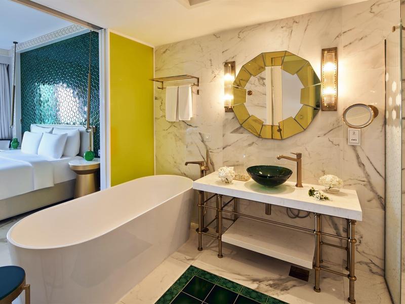 Modern hotel bathroom with freestanding bathtub, green vessel sink, ornate mirror, and marble walls.