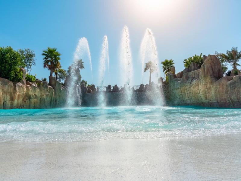 Clear blue water pool with fountains in front of rocky landscape under sunny sky