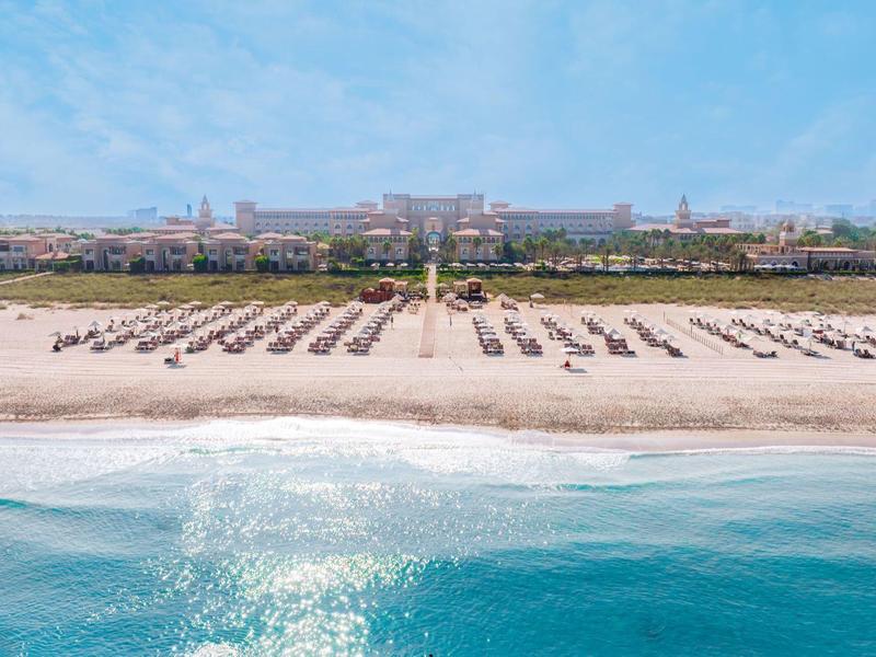 Wide sandy beach with sun loungers in front of a large hotel building under a blue sky.