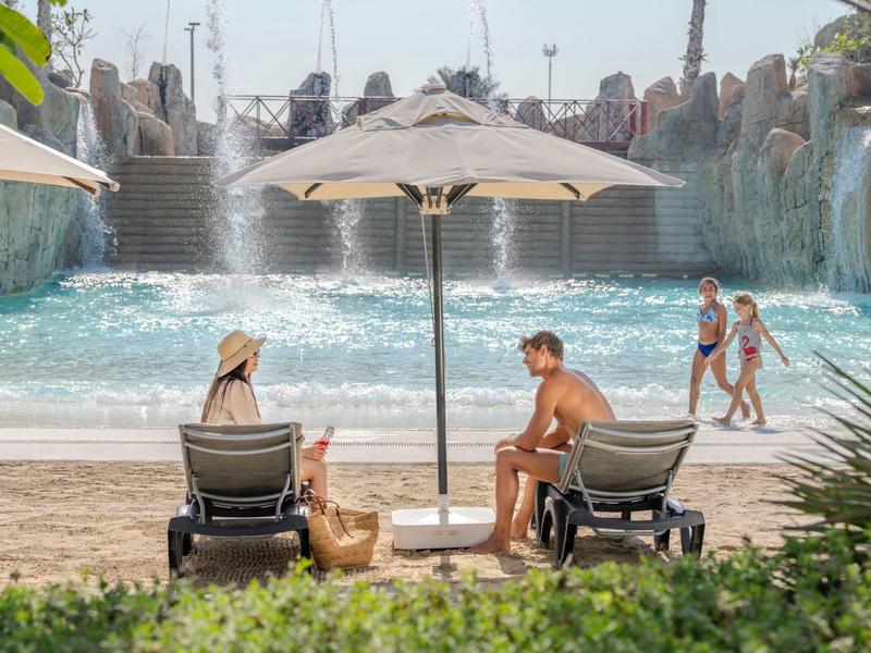 Relaxed people at pool with waterfall and sandy beach under a parasol.