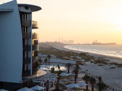 Hotelová budova s balkony u pláže při východu slunce s panoramatem města v pozadí.