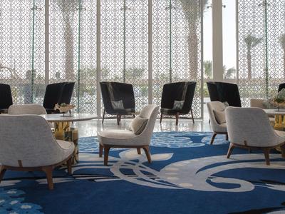 Espace lounge moderne avec fauteuils gris et tapis bleu devant des baies vitrées offrant une vue sur des palmiers.