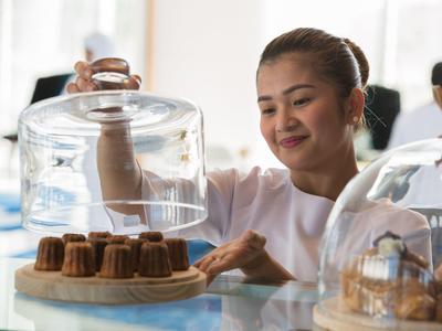 Femme soulève une cloche en verre d'un gâteau dans une boulangerie ou un café lumineux.