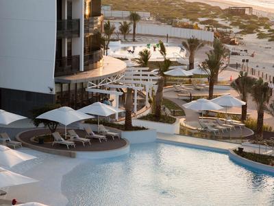 Piscine d'hôtel moderne avec chaises longues, parasols blancs et vue sur la plage au coucher du soleil.