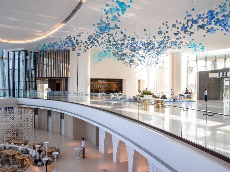 Hall d'hôtel moderne et lumineuse avec une installation décorative bleue au plafond et des espaces de sièges.