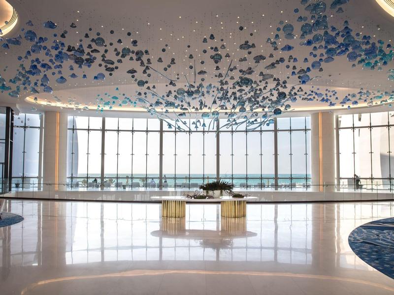 Grand hall d'hôtel lumineux avec grandes fenêtres et installation moderne au plafond composée d'éléments bleus.
