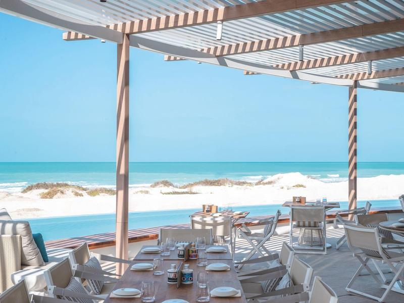 Terrasse ouverte avec tables dressées offrant une vue sur une plage de sable et la mer.