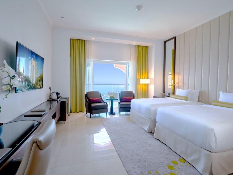 Chambre d'hôtel lumineuse avec deux lits, coin salon et vue sur la mer par une grande fenêtre.