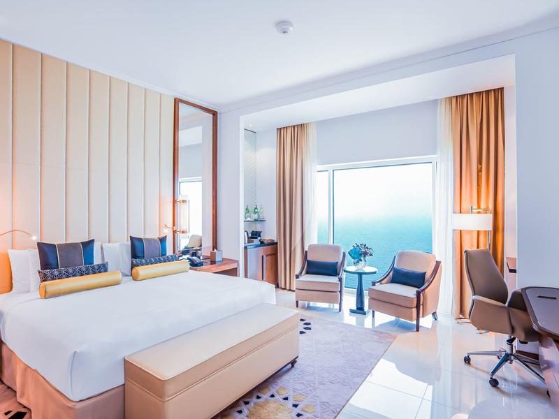 Chambre d'hôtel lumineuse avec grand lit, fauteuils, bureau et vue sur la mer par de grandes fenêtres.