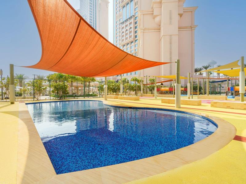 Piscine moderne avec eau bleue et voile d'ombrage orange près d'un grand bâtiment.