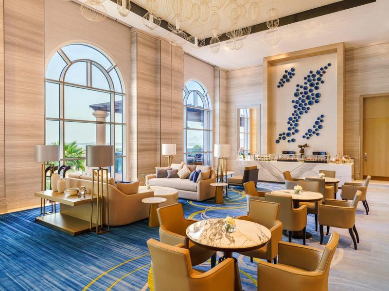 Espace lounge d'hôtel élégant avec tapis bleu, meubles en bois et grandes fenêtres cintrées.