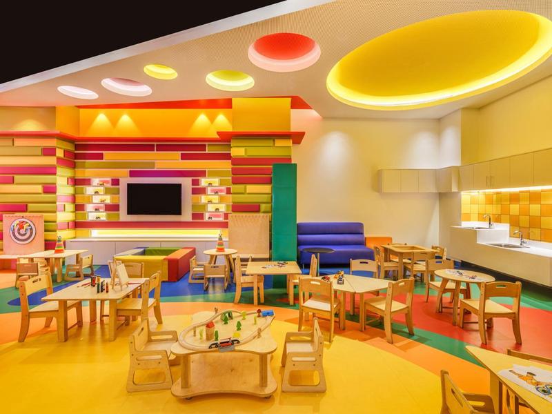 Espace de jeu pour enfants coloré et moderne avec tables, chaises et éclairage au plafond lumineux.
