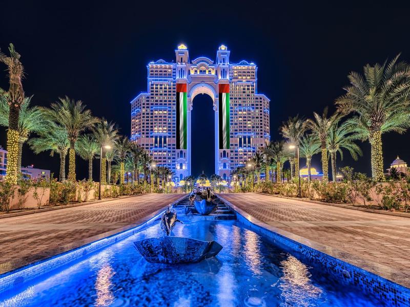 Hôtel moderne éclairé de bleu, palmiers et bassins d'eau la nuit.
