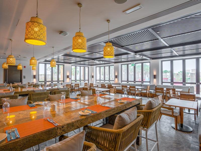Grand espace de restaurant d'hôtel avec tables en bois, chaises en osier et lampes suspendues chaleureuses.