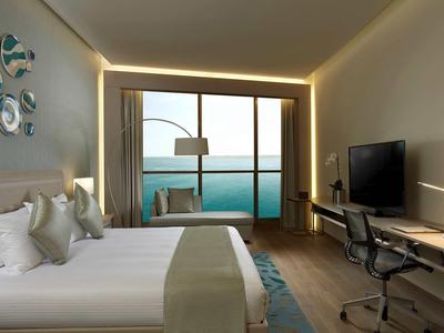 Moderní hotelový pokoj s dvojlůžkem, pracovním stolem a výhledem na moře přes velká okna.