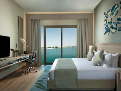 Nowoczesny pokój hotelowy z dużym łóżkiem, biurkiem i widokiem na morze przez szklane drzwi.