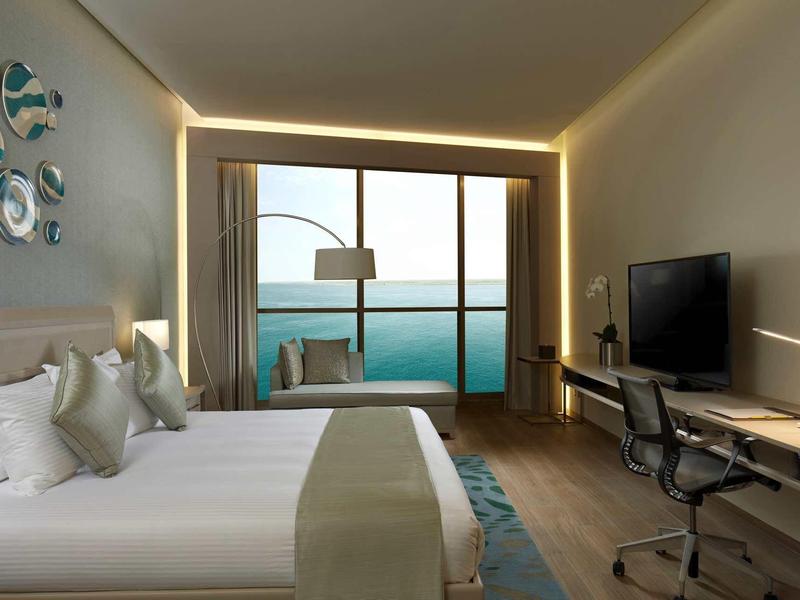 Moderní hotelový pokoj s dvojlůžkem, pracovním stolem a výhledem na moře přes velká okna.