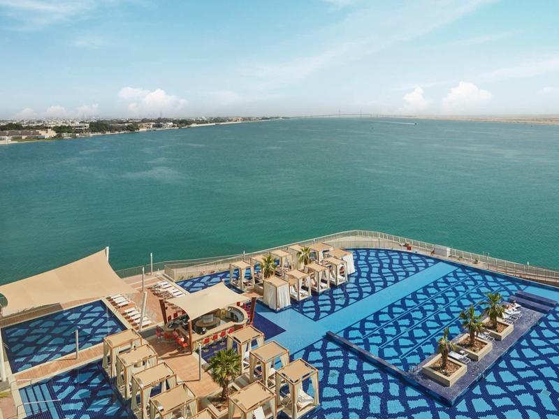 Hotelový bazén s modrou vodou a lehátky na pobřeží s výhledem na moře.