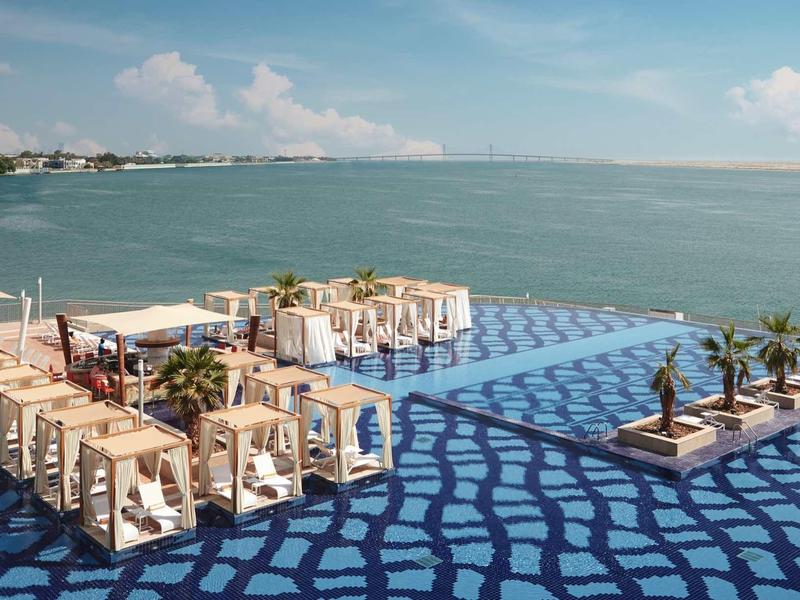 Terasa s židlemi a stoly u vody, modrá vzorovaná podlaha a palmy.