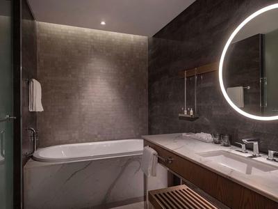 Modernes graues Badezimmer mit eingebauter Badewanne, beleuchtetem Rundspiegel und Holzablage.