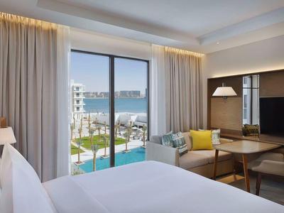 Helles Hotelzimmer mit großem Bett, Sofa, Esstisch, Gardinen, TV und Meerblick durch Glasfenster.