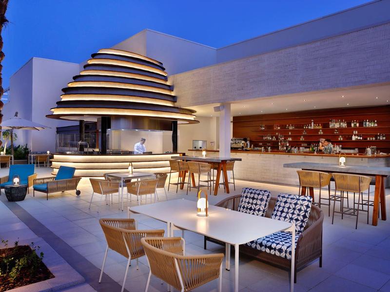 Modern outdoor hotel bar area with lounge chairs and illuminated fireplace at dusk.