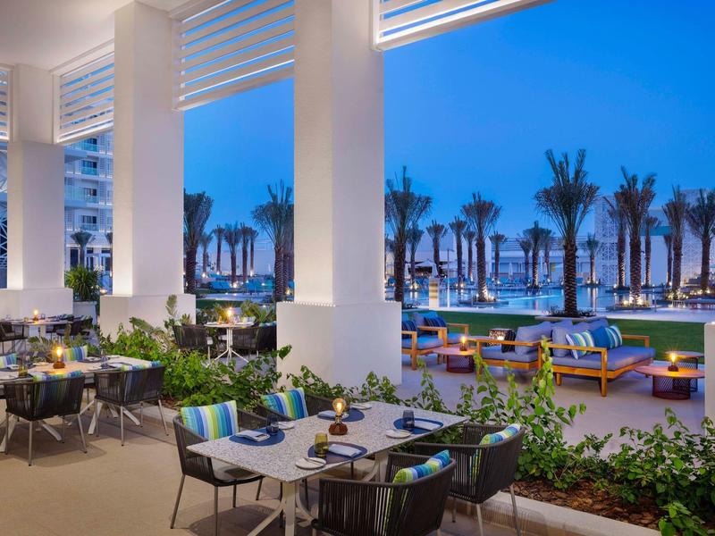 Modern hotel restaurant with outdoor terrace, palm trees, and sea view at twilight.