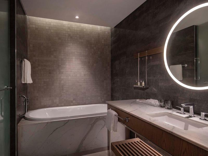 Modern bathroom with illuminated round mirror, double sink, and bathtub in warm gray tones.