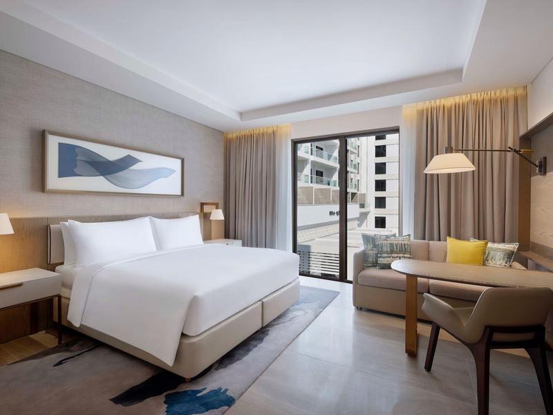 Modern hotel room with double bed, balcony, and seating area overlooking city buildings.