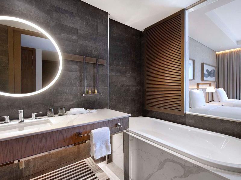 Modern hotel bathroom with illuminated round mirror, bathtub, and view into the bedroom.
