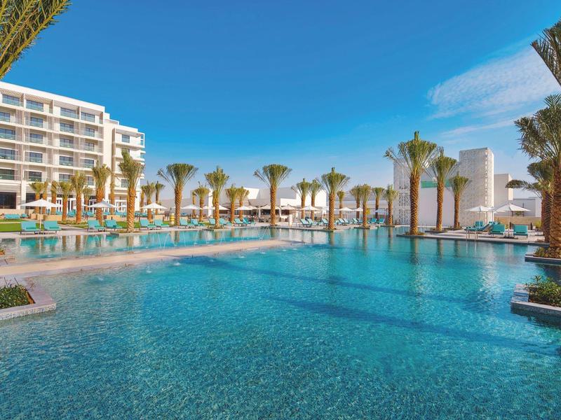 Large outdoor pool with palm trees and modern hotel building under clear sky.