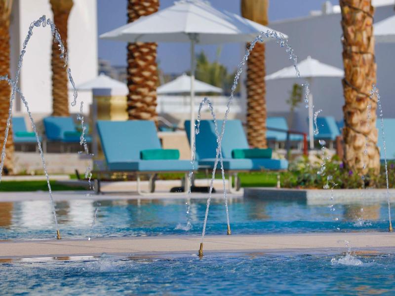 Modern hotel pool with blue loungers, white umbrellas, and palm trees in the background.