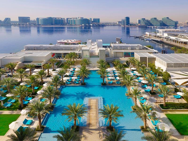 Luxury hotel complex with large pools, palm trees, and sea view with modern buildings.