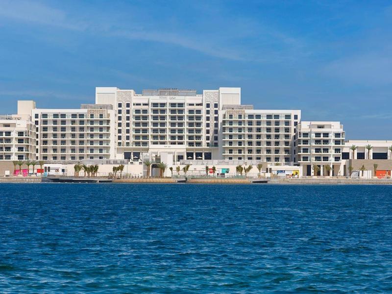 Large white hotel building by the sea with blue sky and water in the foreground.