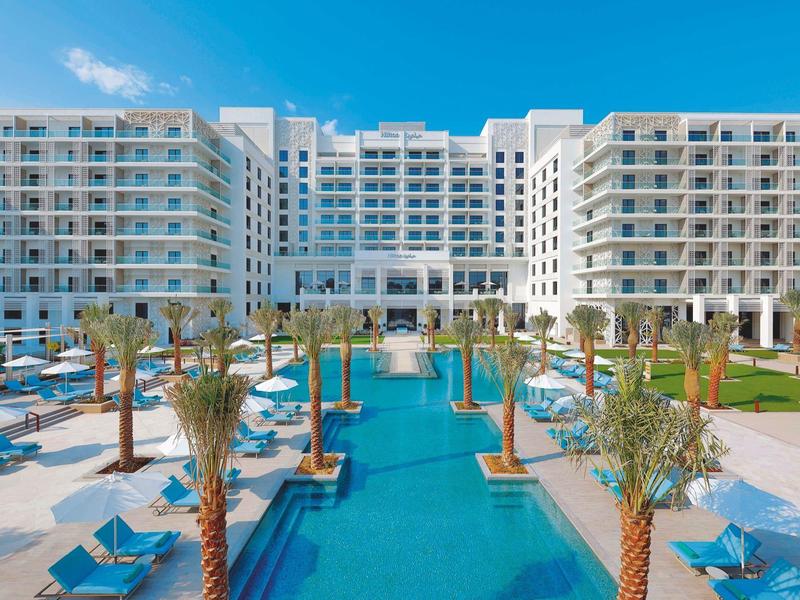 Modern hotel complex with large pool and palm trees under a blue sky