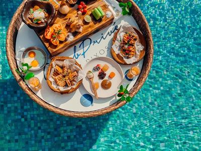 Bandeja flotante con varios platos de desayuno sobre agua azul de piscina.