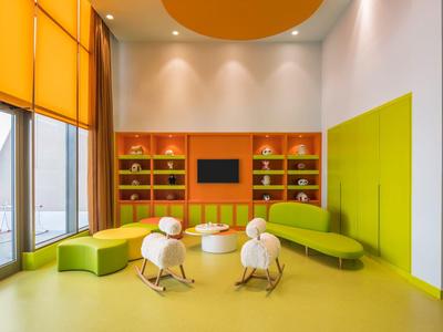 Sala de espera moderna de hotel con sofás verdes, sillas para niños y grandes ventanas con persianas naranjas.