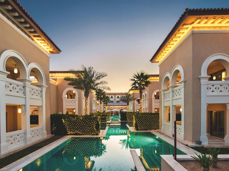 Vista al atardecer de un lujoso hotel con piscina central y palmeras entre edificios con arcos blancos.