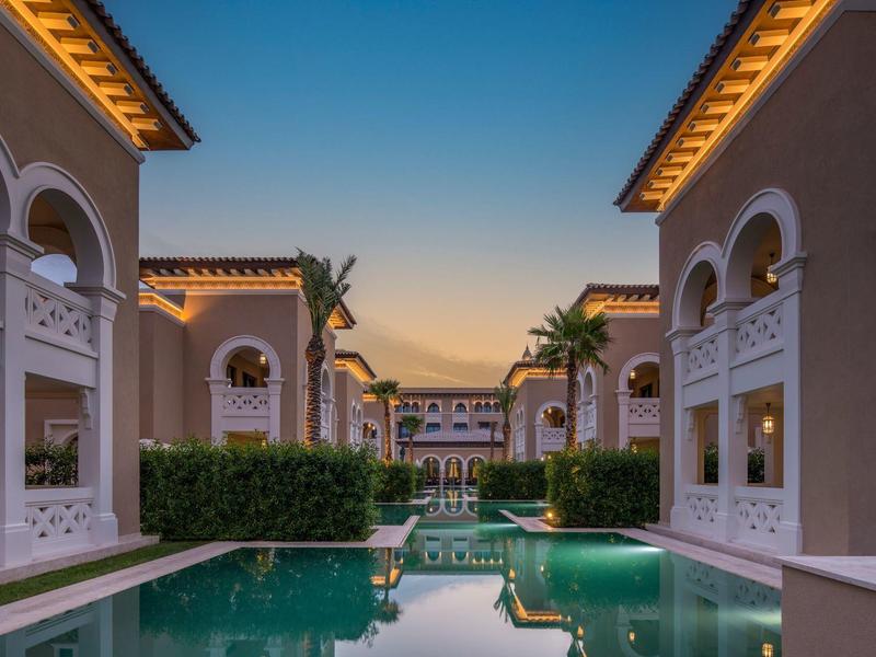 Un hotel de lujo con edificios iluminados, palmeras y una piscina azul claro al atardecer.