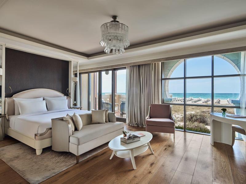 Elegante habitación de hotel con vista al mar, cama grande, sofá, sillón y suelo de madera.