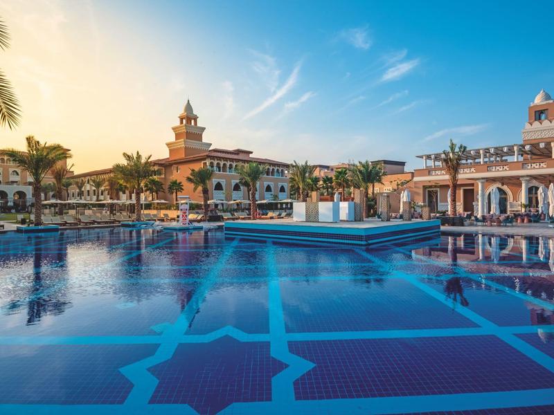 Gran piscina con agua azul frente a un edificio histórico de hotel al atardecer.
