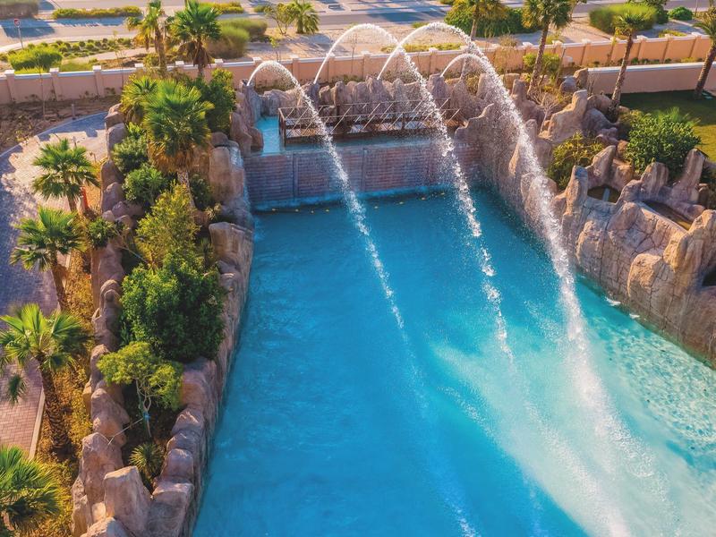 Piscina azul con fuentes de agua, rodeada de palmeras y estructuras de piedra en un hotel.