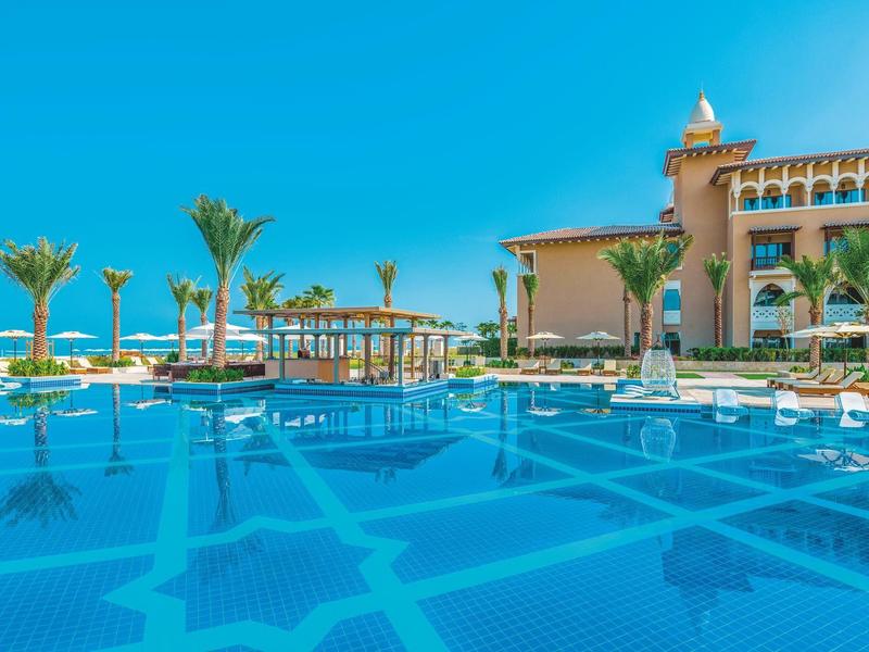 Gran piscina del hotel con suelo azul decorado, palmeras y tumbonas bajo cielo despejado.
