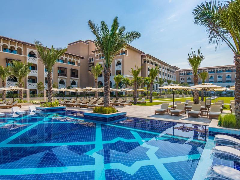 Hotel de lujo con piscina azul, palmeras y tumbonas bajo un cielo despejado.