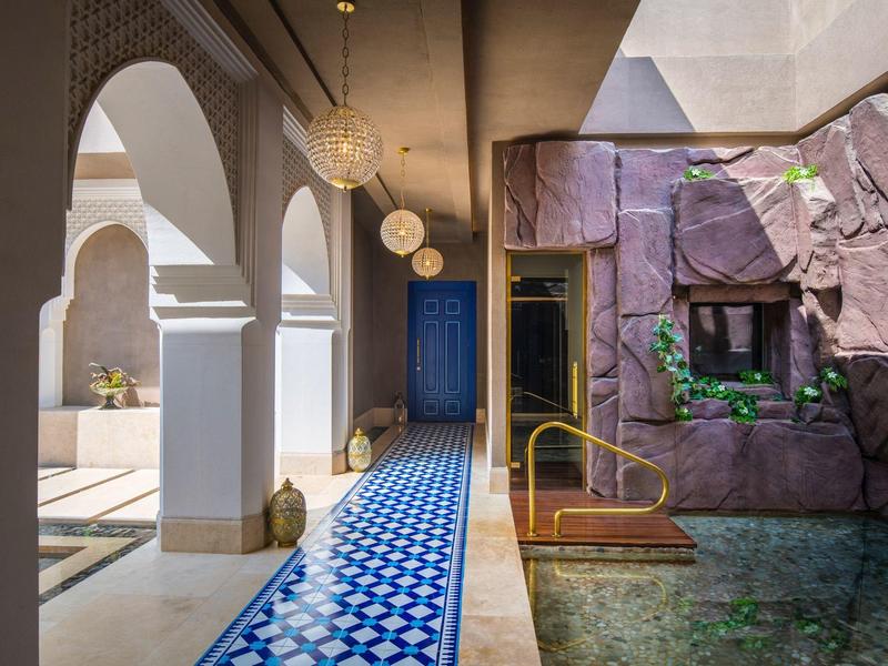 Pasillo con suelo de azulejos azules, arcos blancos y piscina junto a muro morado en hotel