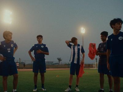 Vijf jongeren in sportoutfits staan op een verlicht voetbalveld bij schemering.